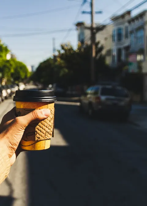Hand hält einen Coffee-to-go-Becher in einer sonnigen Straße